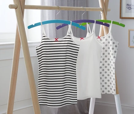 Trois maillots de corps pour filles, un rayé noir et blanc, un blanc et un à pois gris, suspendus à des cintres sur un portant en bois.