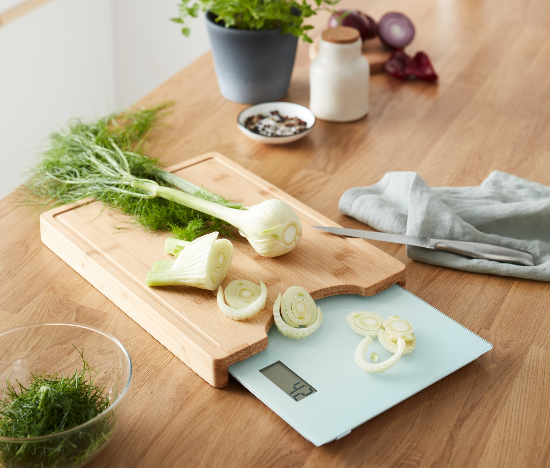 Sur une surface en bois se trouve une planche à découper avec balance de cuisine amovible sur laquelle est posé du fenouil coupé.