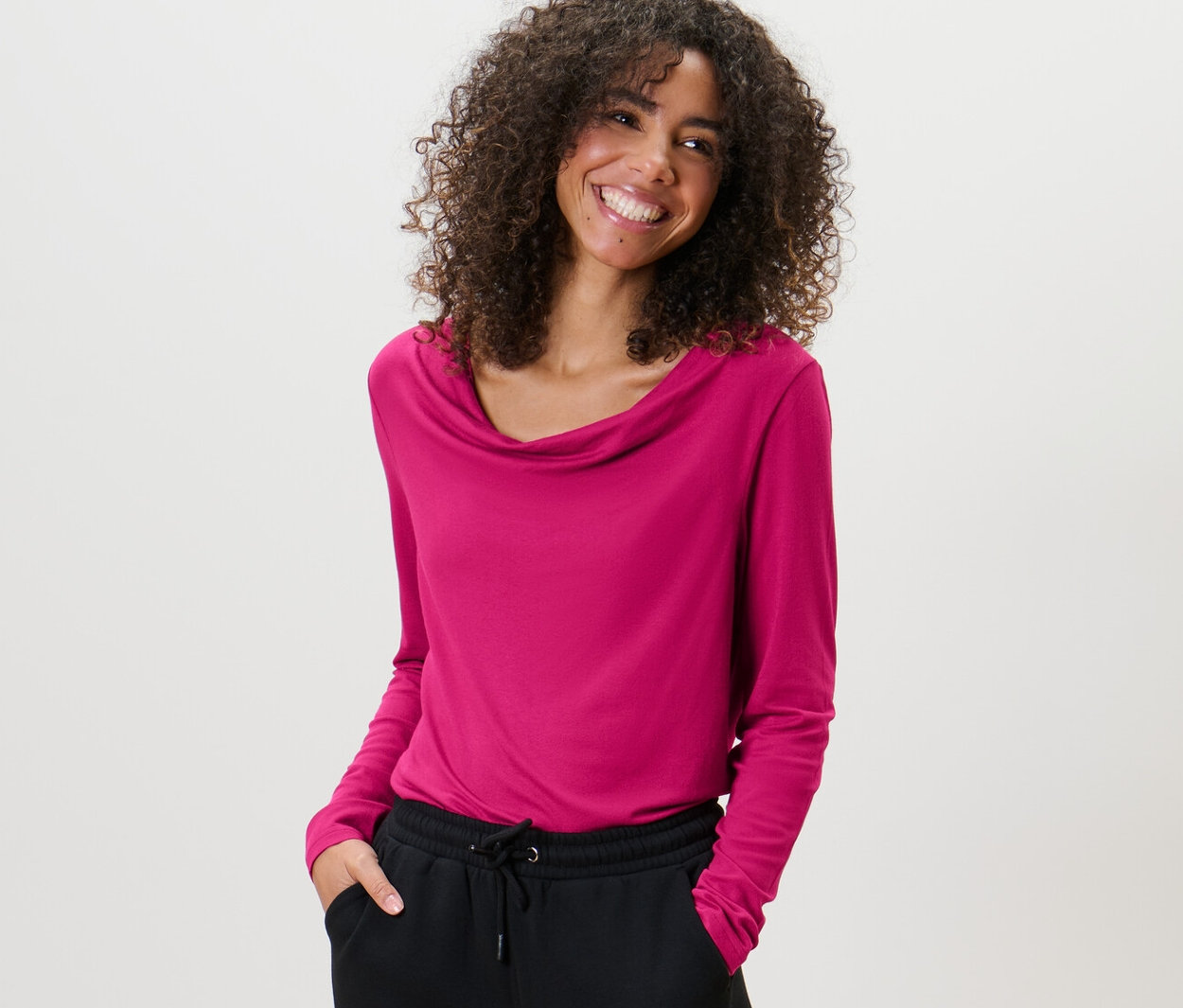 Femme aux cheveux bouclés portant une blouse avec encolure en cascade et un pantalon noir.