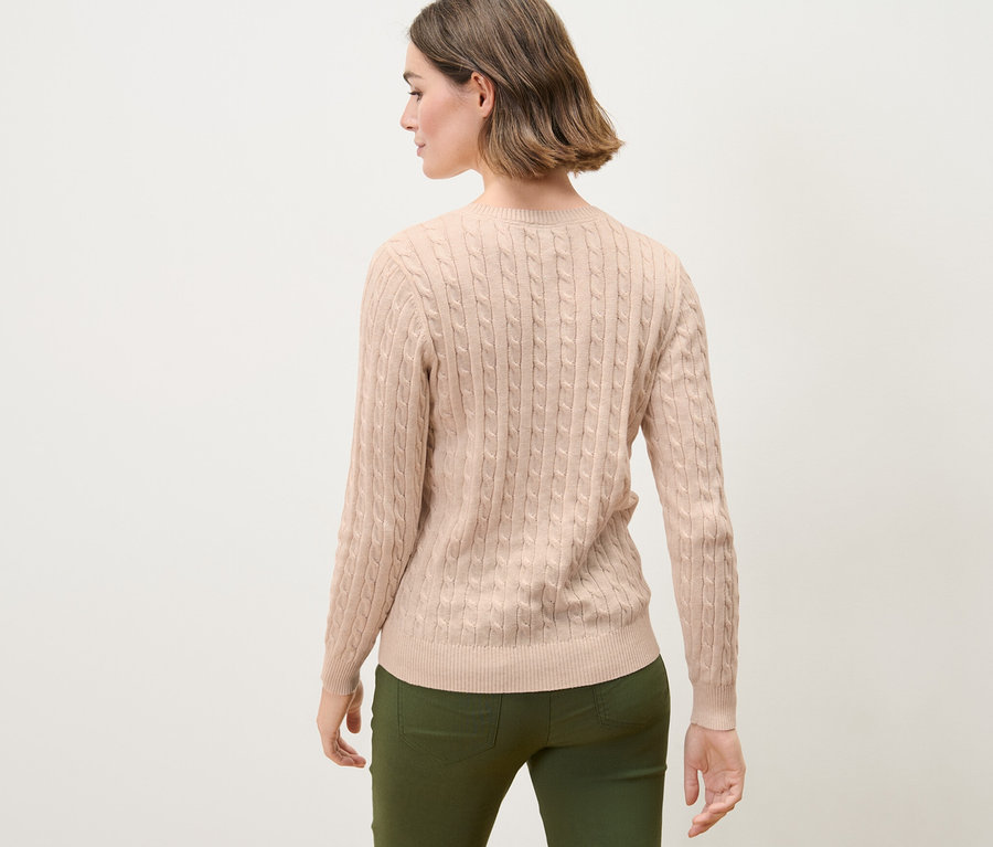 Une femme aux cheveux bruns pose dos à la caméra et porte un pull en maille torsadée de couleur crème.