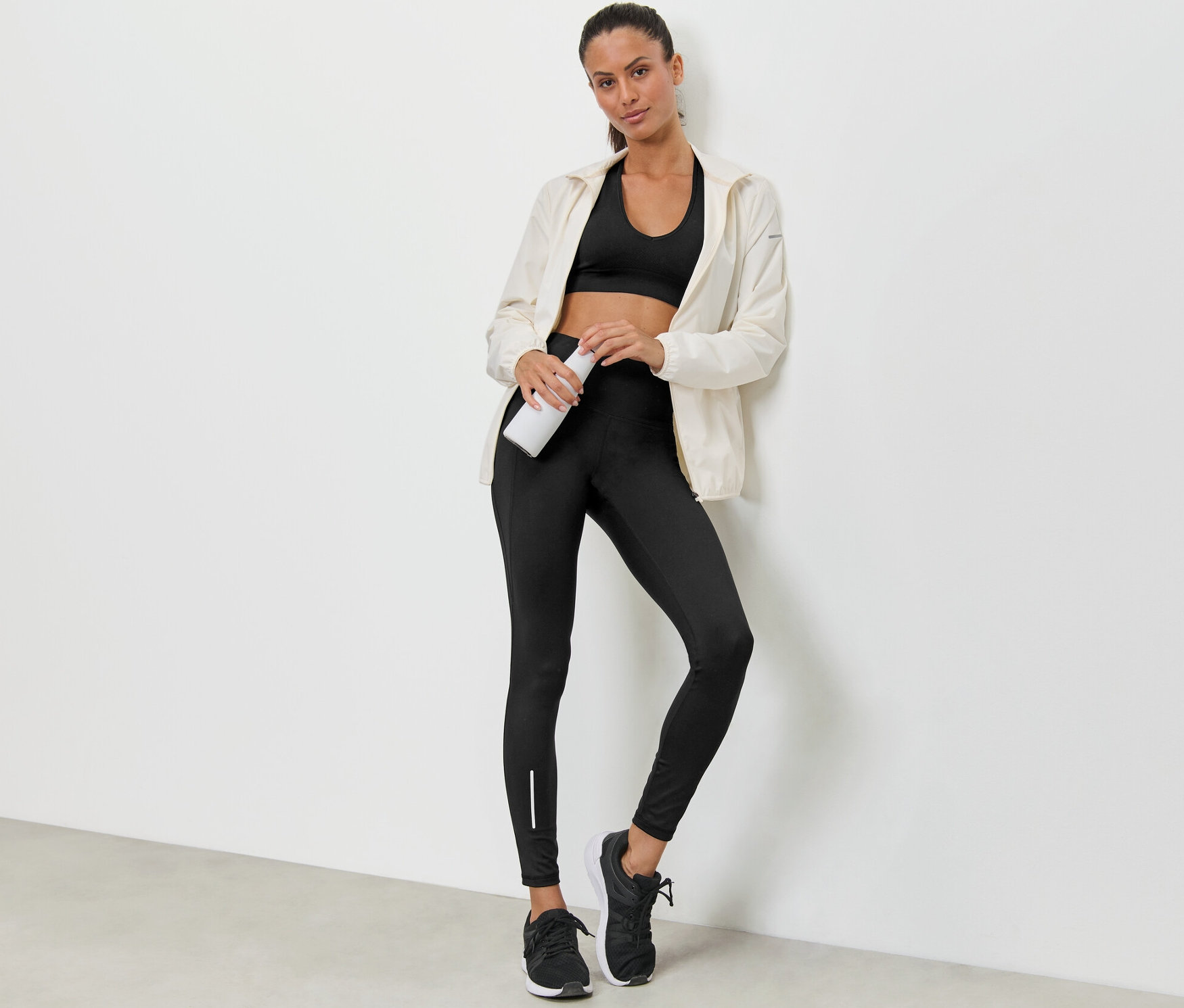 Une femme appuyée contre un mur tenant une bouteille d'eau, porte un soutien-gorge de sport, une veste et un legging de sport déperlant.