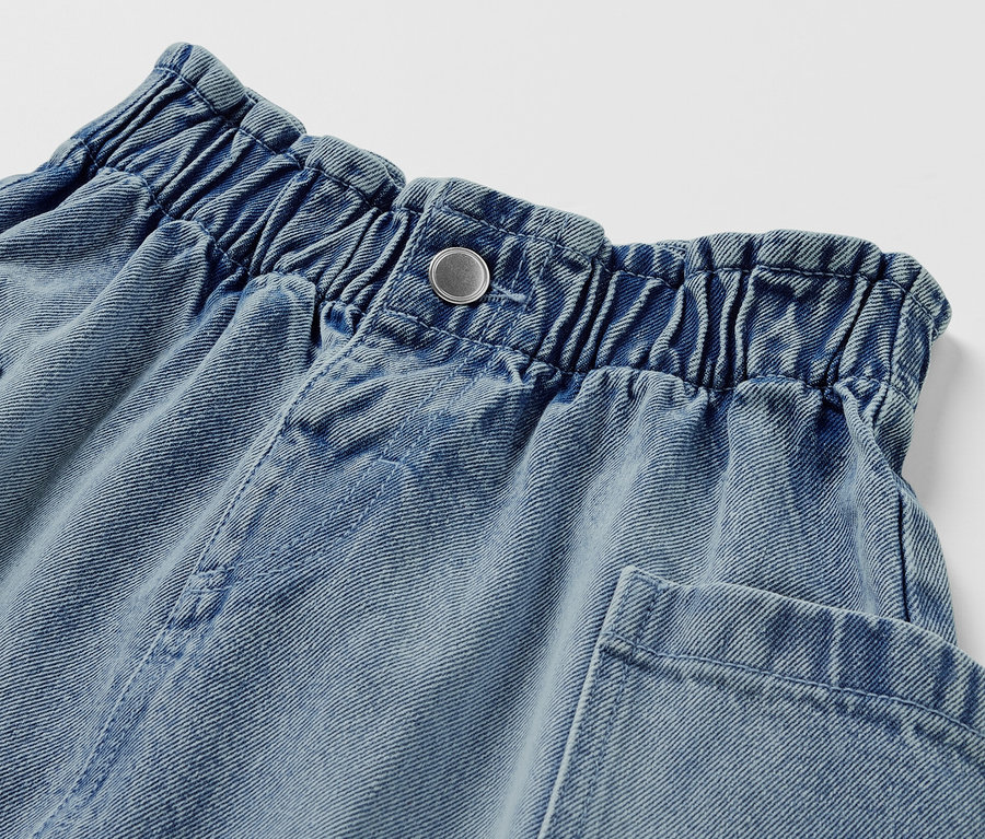 Détail d'une jupe paper bag en jeans pour enfant avec fermeture à bouton et poches.