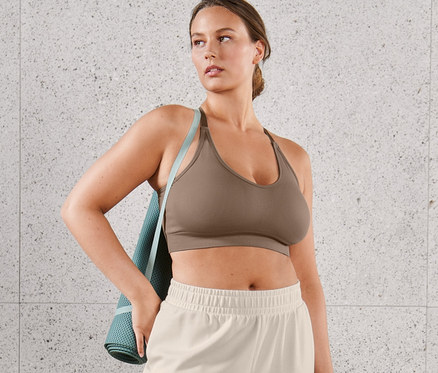 Une femme porte une brassière de sport sans coutures marron et un pantalon molletonné très doux. Un tapis de yoga est drapé sur son épaule.