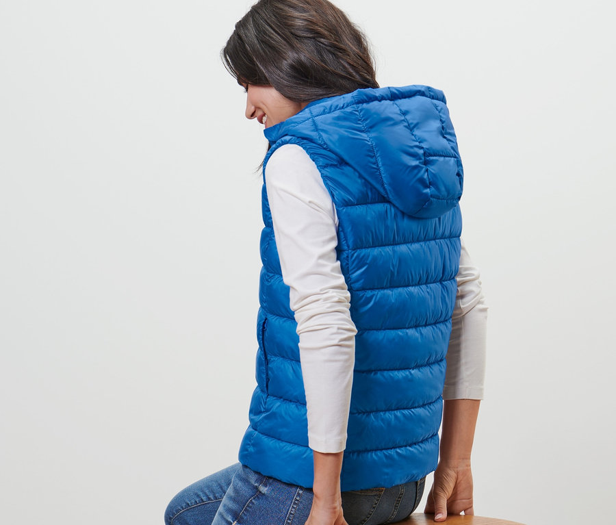 Femme assise sur un tabouret en bois portant un gilet matelassé à capuche bleu.