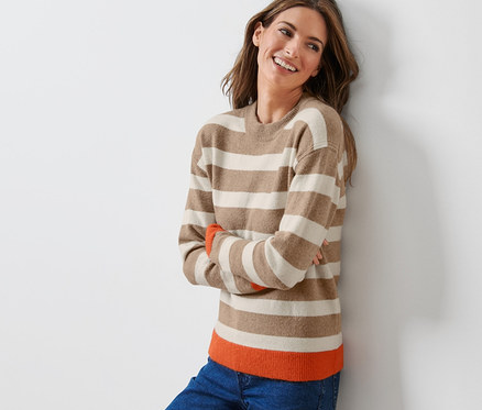 Une femme souriante, adossée contre un mur, porte un pull en maille à rayures.