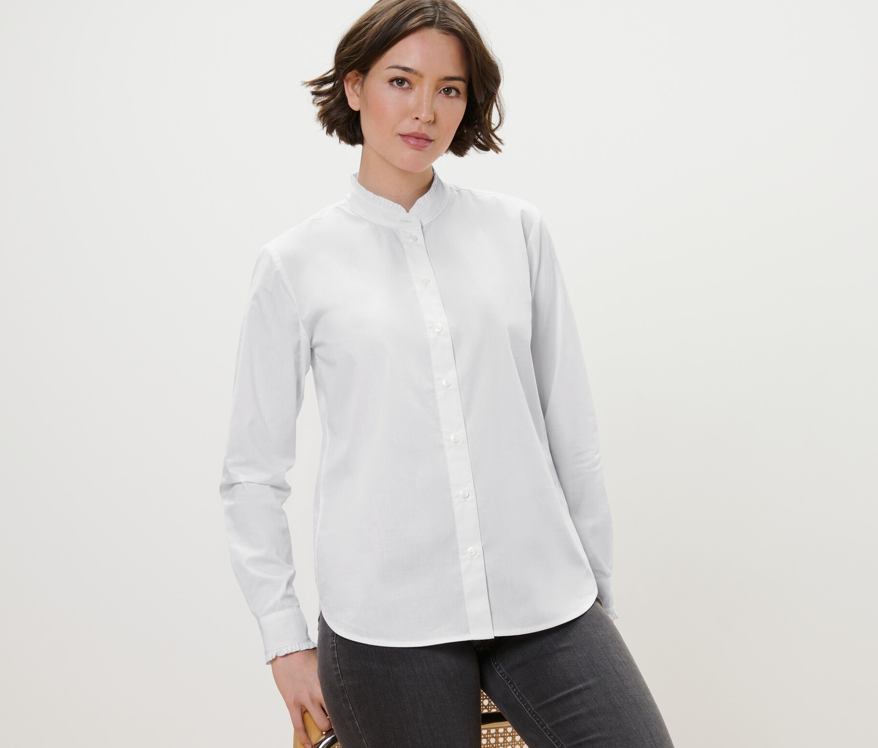 Une femme pose sur une chaise et porte un chemisier blanc avec des détails ruchés et un jean gris.