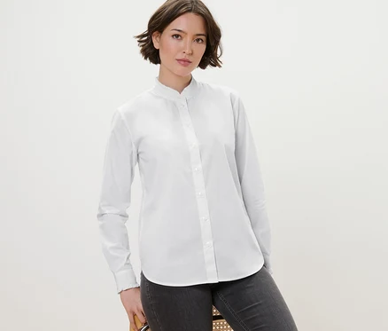Une femme pose sur une chaise et porte un chemisier blanc avec des détails ruchés et un jean gris.