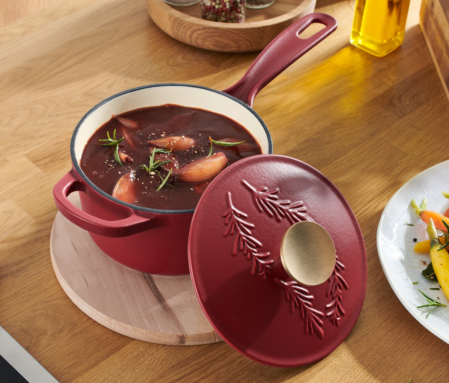 Une casserole en fonte rouge avec couvercle sur une table en bois à côté d'une assiette de nourriture.