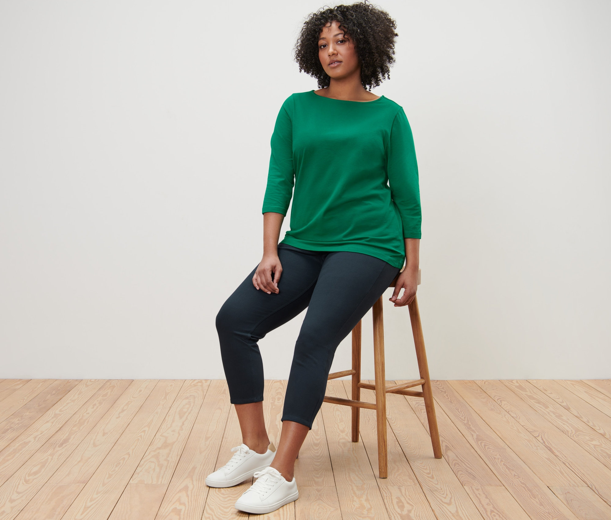 Une femme est assise sur un tabouret et porte un t-shirt vert à manches 3/4 et un jegging 7/8.