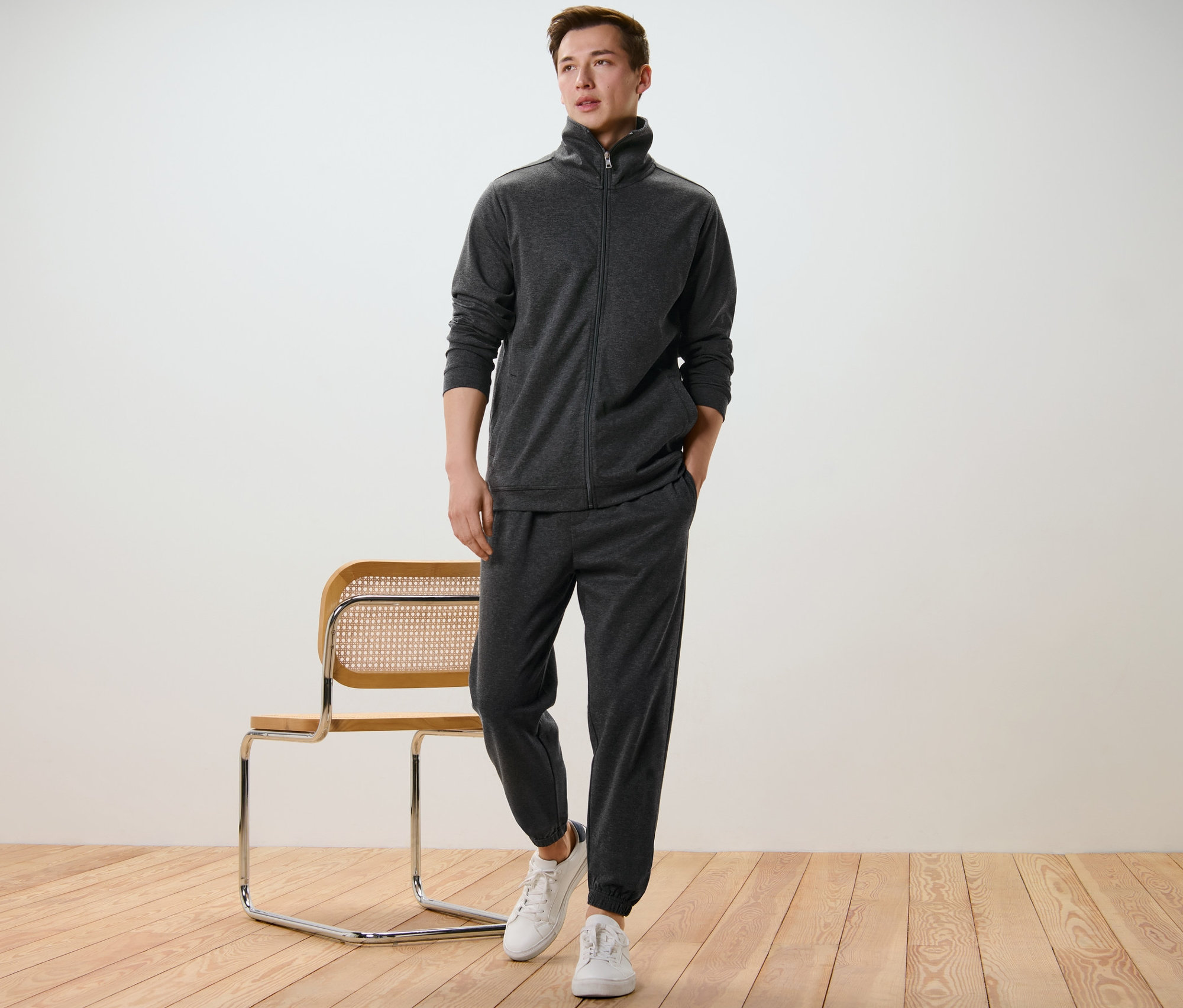 Un homme pose en survêtement d’intérieur et baskets blanches à côté d’une chaise.