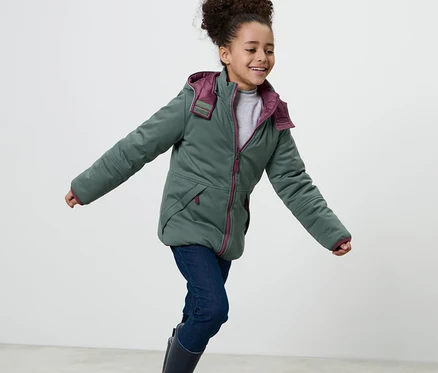 Fillette souriante portant une veste matelassée et softshell 2 en 1 pour enfant avec capuche, un jean et des bottes en caoutchouc.