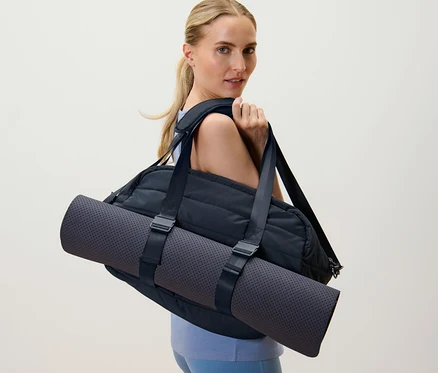 Une femme blonde porte un sac de sport noir et un tapis de yoga sur l'épaule.