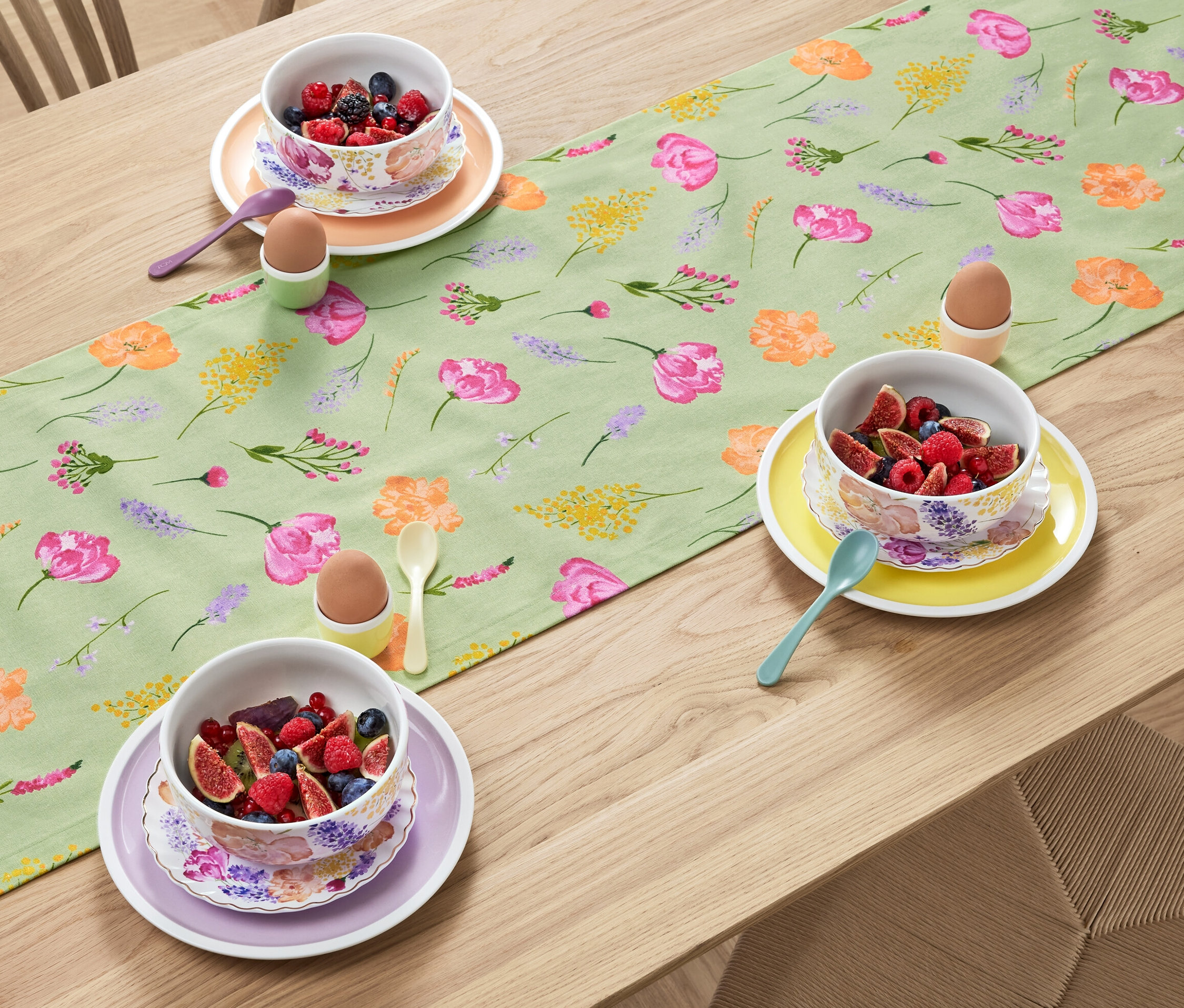 Sur une table en bois, un chemin de table sauge à imprimé floral est disposé avec des bols de fruits, des œufs dans des coquetiers et des cuillères.