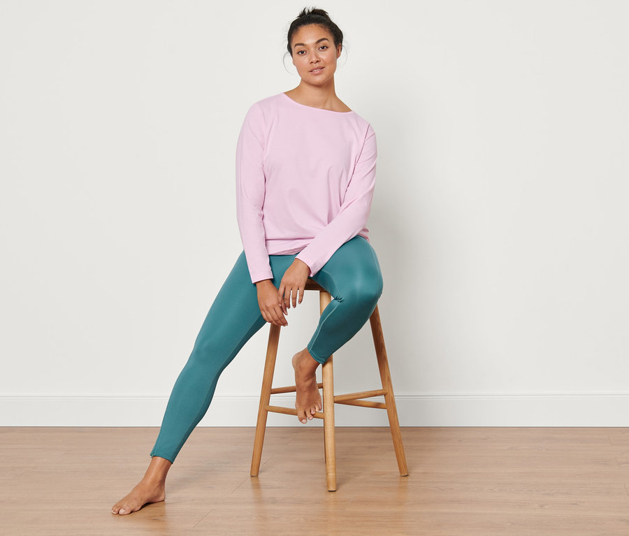 Une femme est assise sur un tabouret et porte un legging de sport modelant vert et un t-shirt de sport manches longues rose clair.