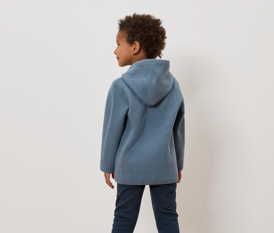 Un garçon aux cheveux bouclés porte un sweat à capuche bleu et un pantalon bleu foncé.