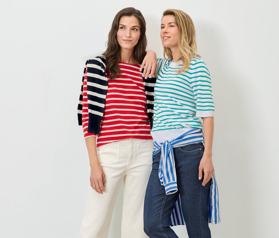Deux femmes posent sur un fond blanc. L'une porte un pull en maille fine rayé turquoise et un jean, l'autre un pull rayé rouge et blanc et un pantalon blanc.