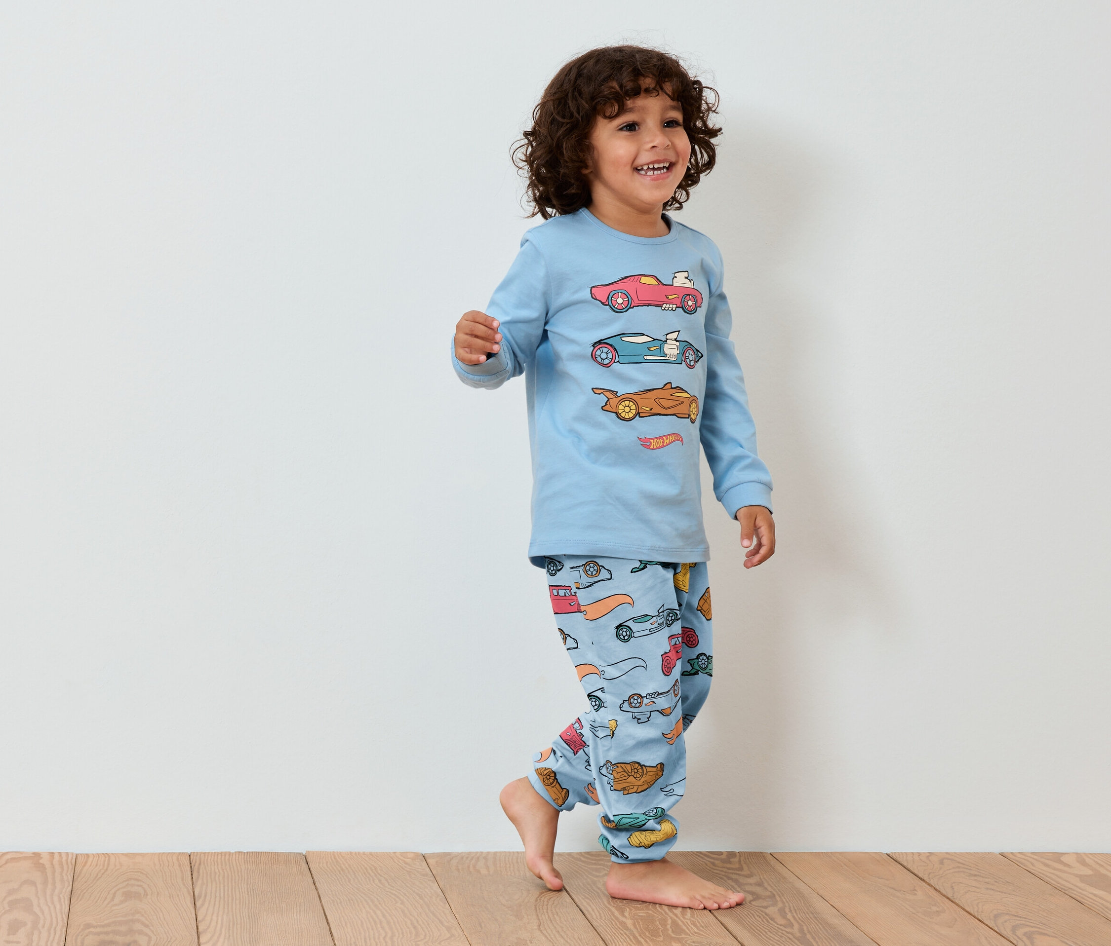 Petit garçon souriant en pyjama bleu avec des voitures marchant sur un plancher en bois.