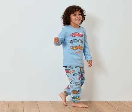 Petit garçon souriant en pyjama bleu avec des voitures marchant sur un plancher en bois.