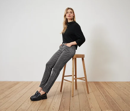 Une femme blonde est assise sur un tabouret en bois, portant un t-shirt noir à manches longues, un pantalon de velours côtelé gris et des mocassins noirs.