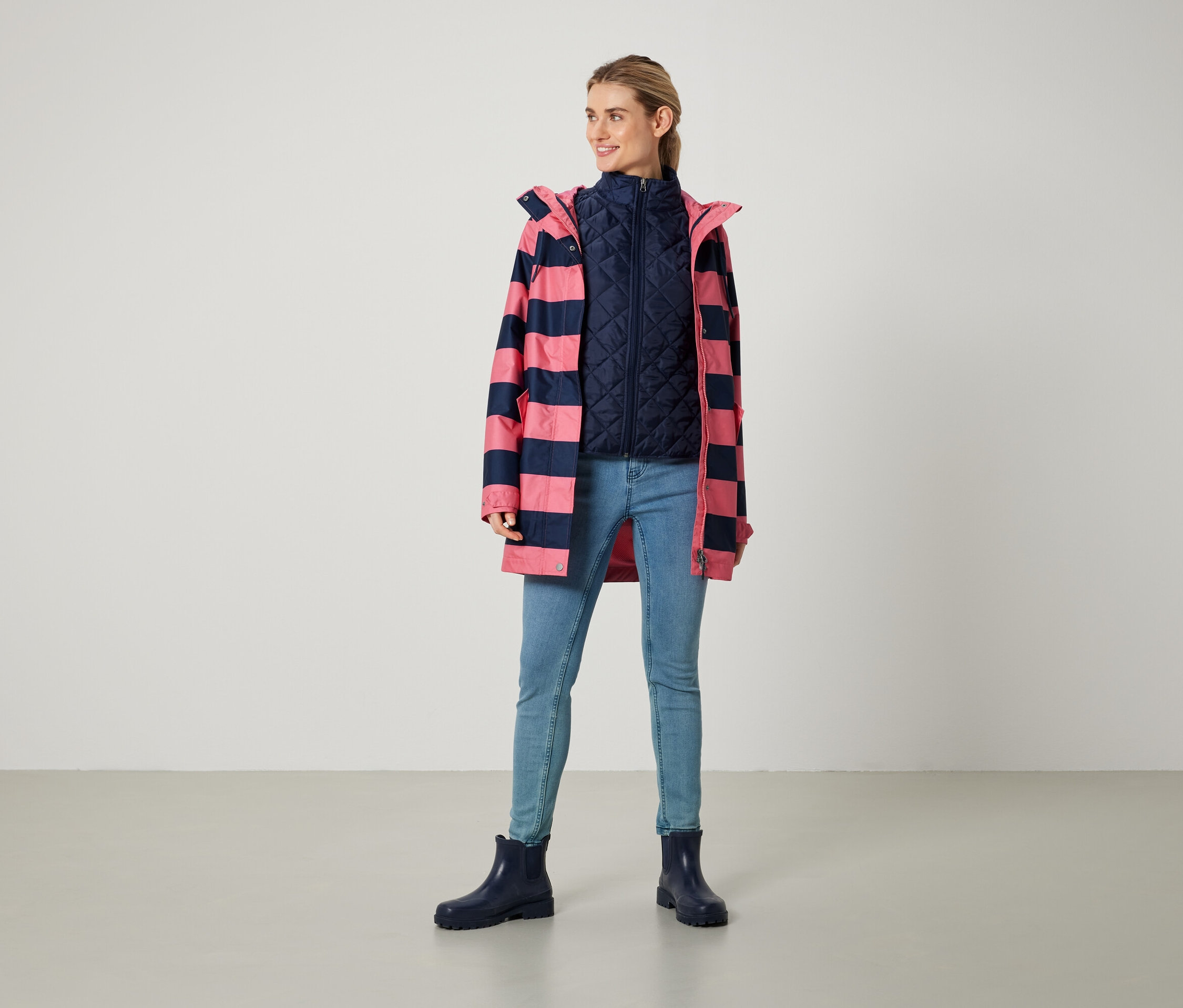 Une femme debout porte un manteau rayé rose et marine, un gilet marine, un jean et des bottes bleues.