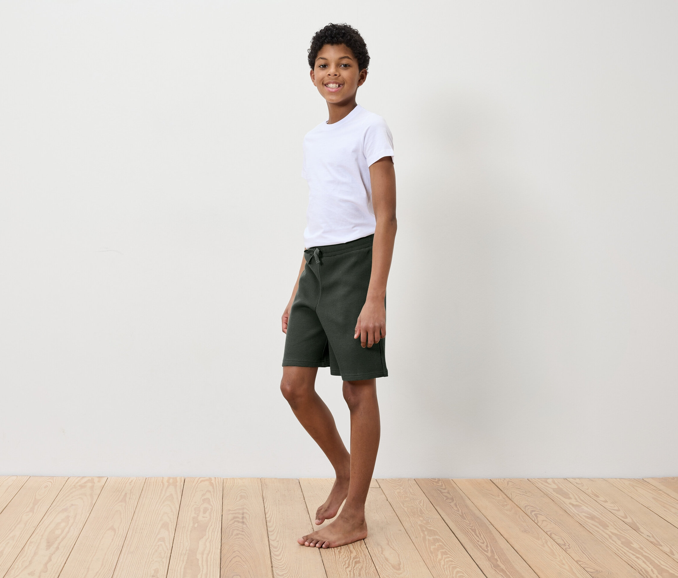 Un garçon en t-shirt blanc et short vert debout sur un plancher en bois devant un mur blanc.