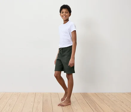 Un garçon en t-shirt blanc et short vert debout sur un plancher en bois devant un mur blanc.