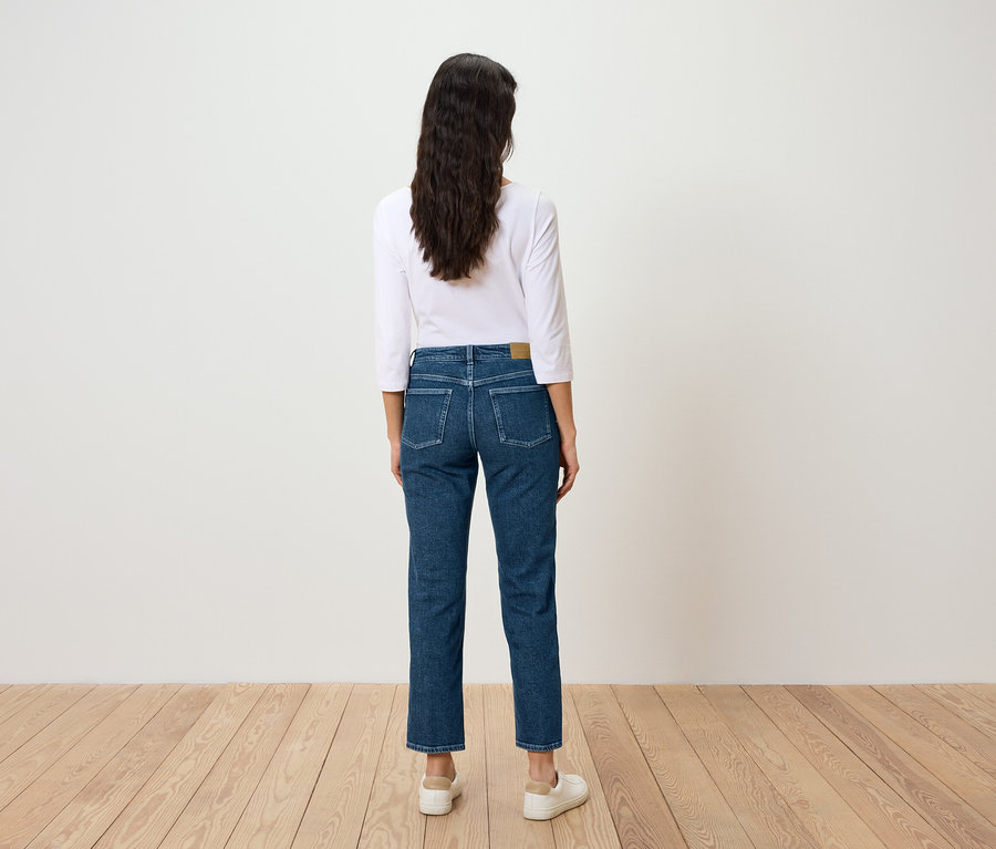 Une femme aux longs cheveux foncés est vue de dos, portant un jean bleu et un t-shirt blanc.