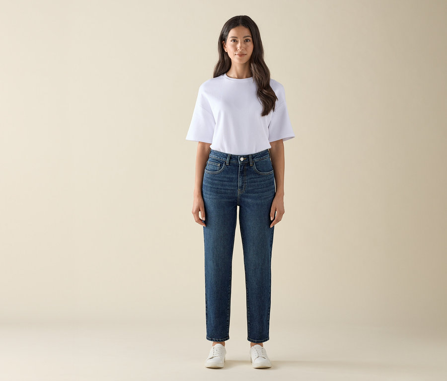 Une femme debout portant un Mom Jeans – Coupe « Nyla » bleu foncé, un t-shirt blanc et des baskets blanches.