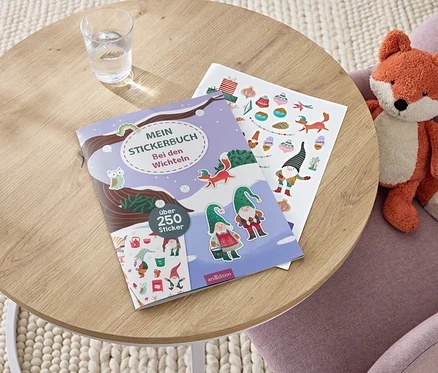 Sur une table se trouvent un livre d'autocollants, une feuille d'autocollants et un verre d'eau. Sur une chaise est assis un renard en peluche.