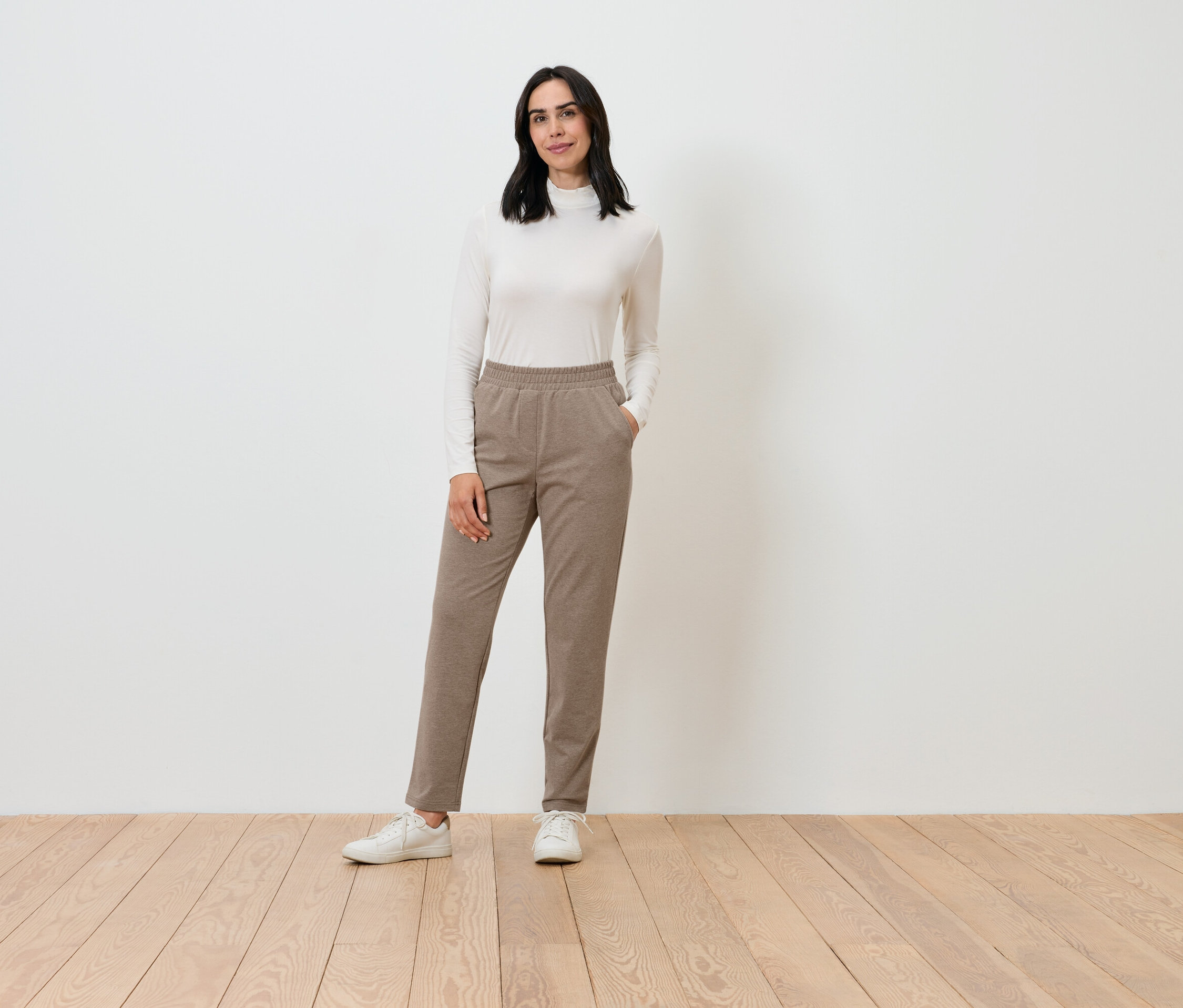 Une femme pose, portant un col roulé crème et un pantalon marron.