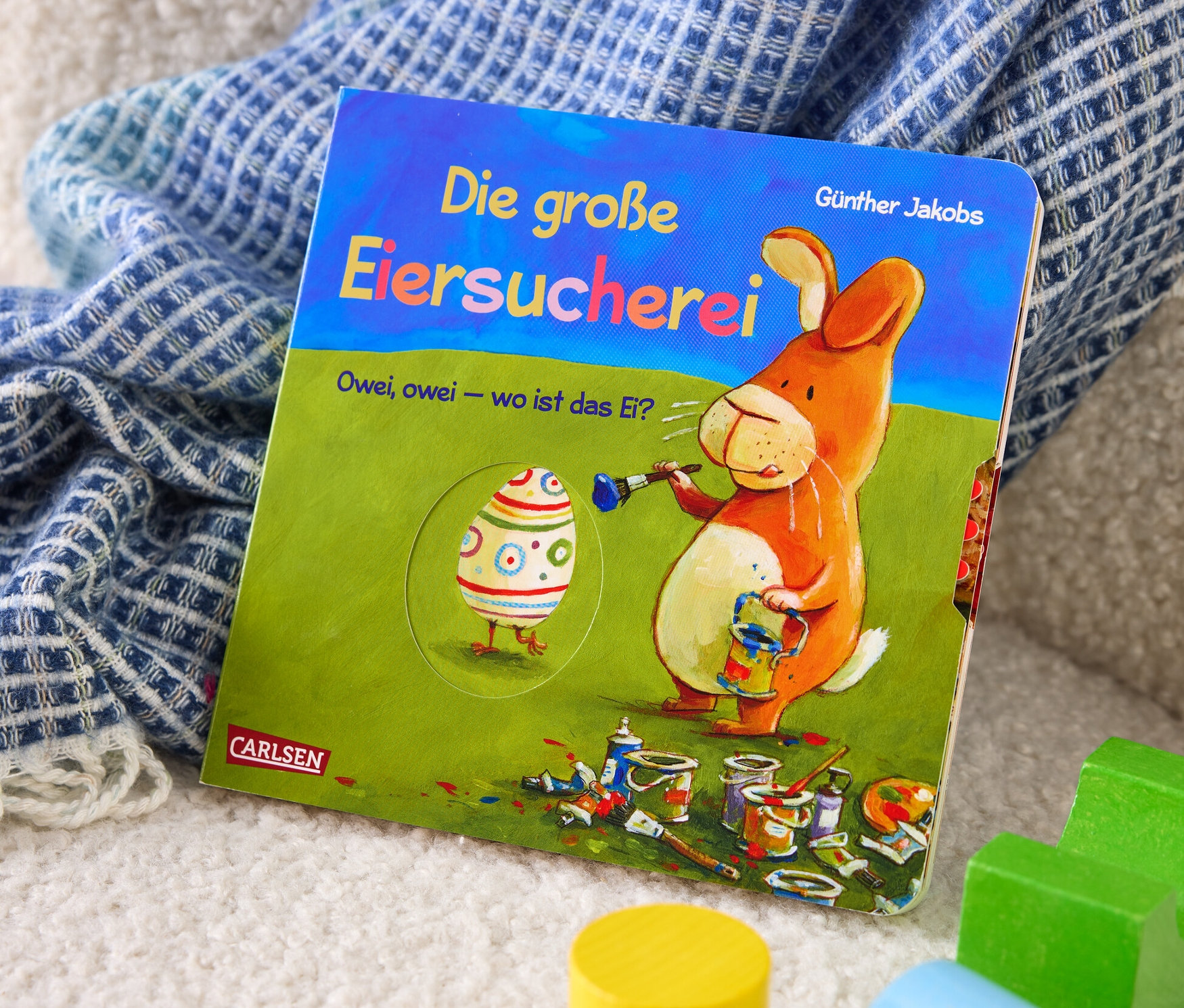 Le livre « La grande chasse aux œufs » (en allemand) est posé sur un jeté à côté de cubes en bois.