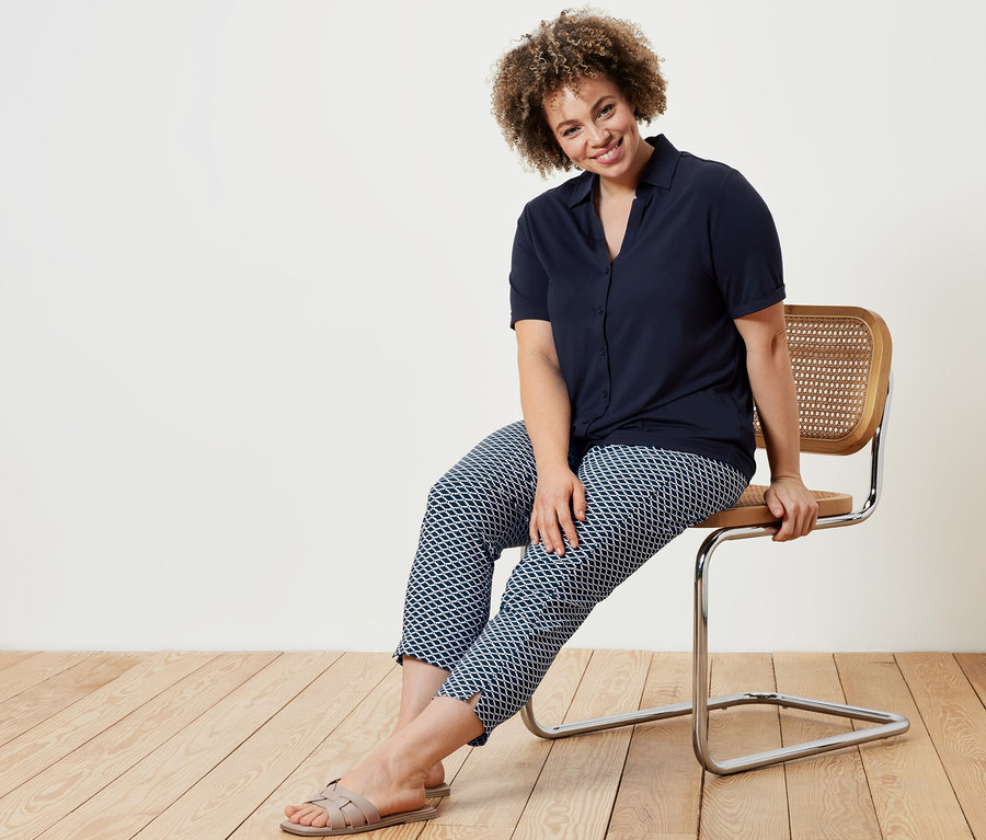 Une femme assise sur une chaise porte un chemisier bleu foncé avec une patte de boutonnage et un pantalon stretch imprimé 7/8.