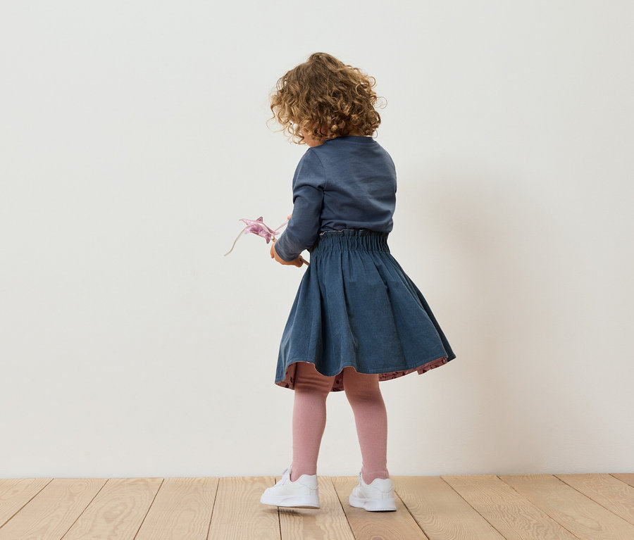 Une fillette aux cheveux bouclés, vue de dos, tient un jouet, porte une jupe bleue et des baskets blanches.