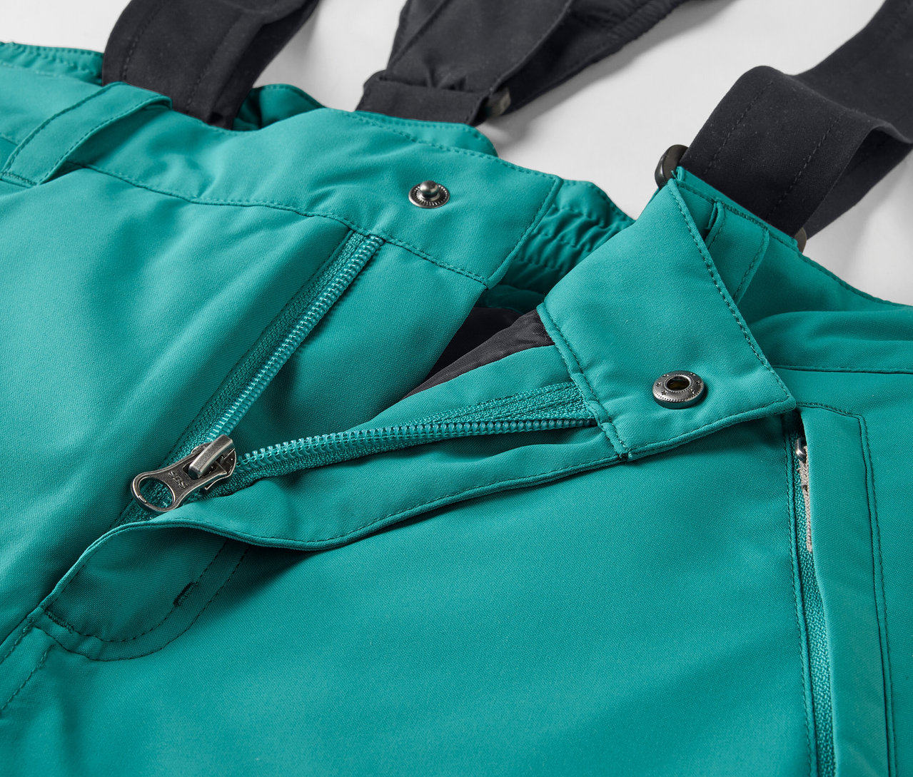 Détail d'un pantalon de ski vert avec des bretelles noires et une fermeture éclair ouverte.