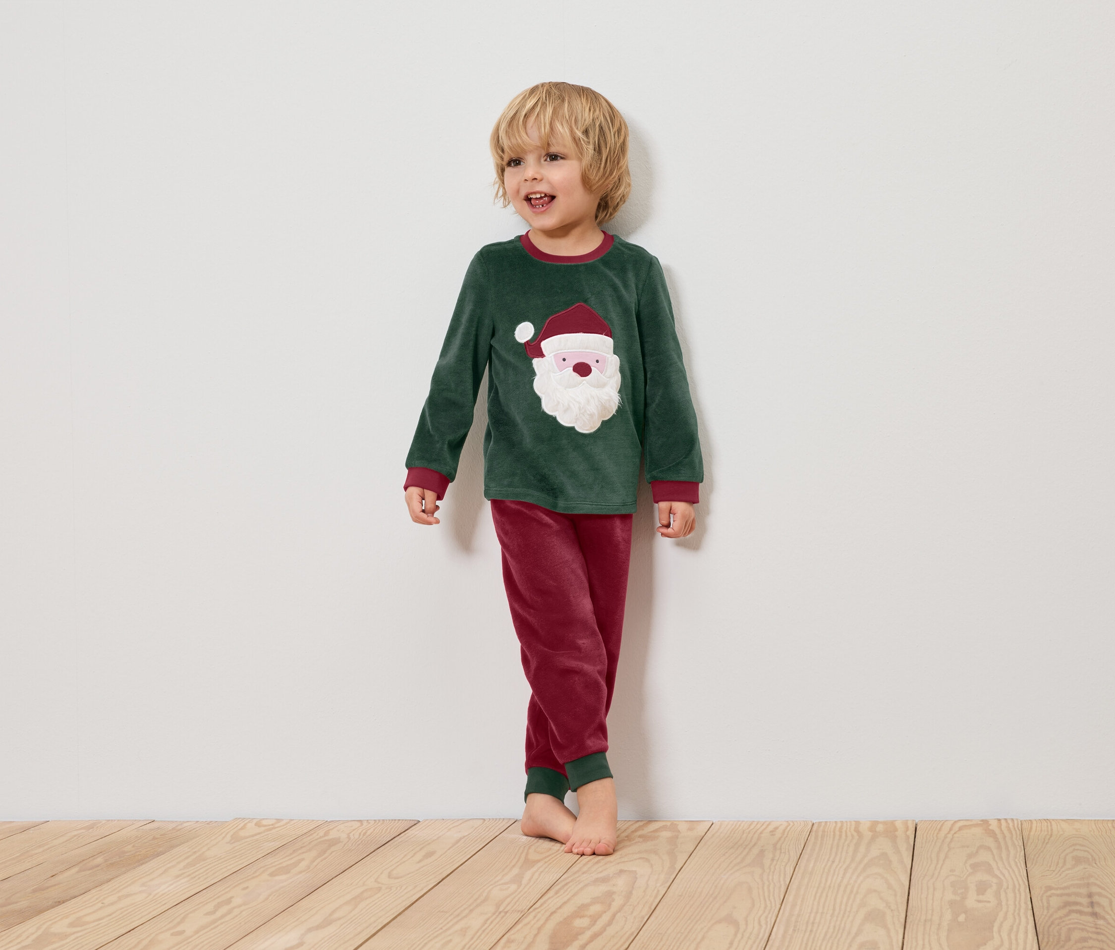 Un garçon en pyjama de Noël vert avec un motif de Père Noël se tient sur un plancher en bois devant un mur blanc.