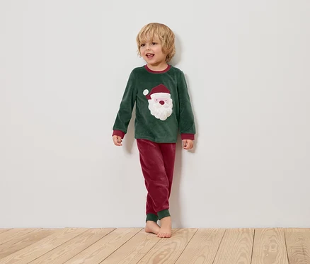 Un garçon en pyjama de Noël vert avec un motif de Père Noël se tient sur un plancher en bois devant un mur blanc.