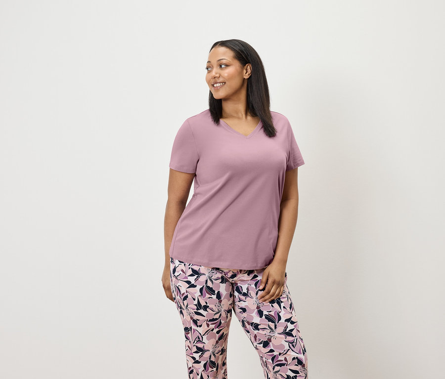 Femme en t-shirt lilas à manches courtes et pantalon à fleurs regardant vers la droite.