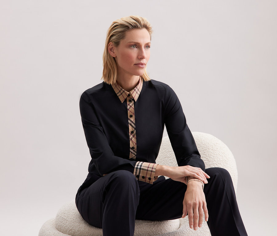 Une femme blonde est assise sur une chaise blanche et porte des vêtements noirs avec des motifs à carreaux.