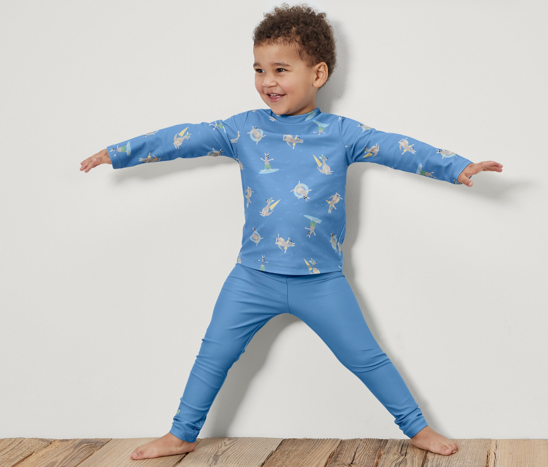 Un garçon se tient debout, les bras écartés, et porte un legging de bain pour enfant avec un indice de protection UV 80.