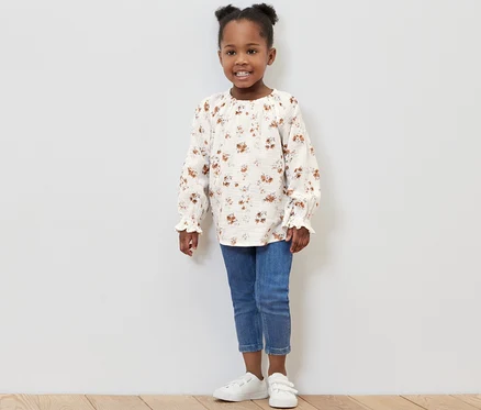 Petite fille souriante posant en studio portant une blouse en mousseline pour enfant, un jean et des baskets.