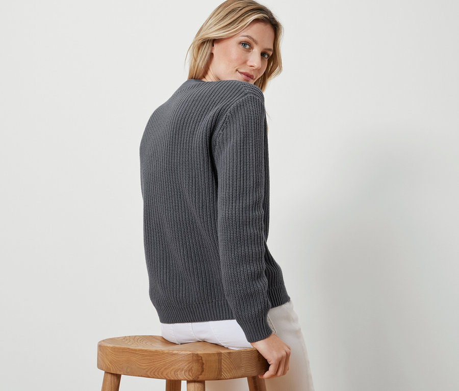 Une femme blonde est assise de côté sur un tabouret en bois et regarde par-dessus son épaule, portant un cardigan anthracite en grosse maille.