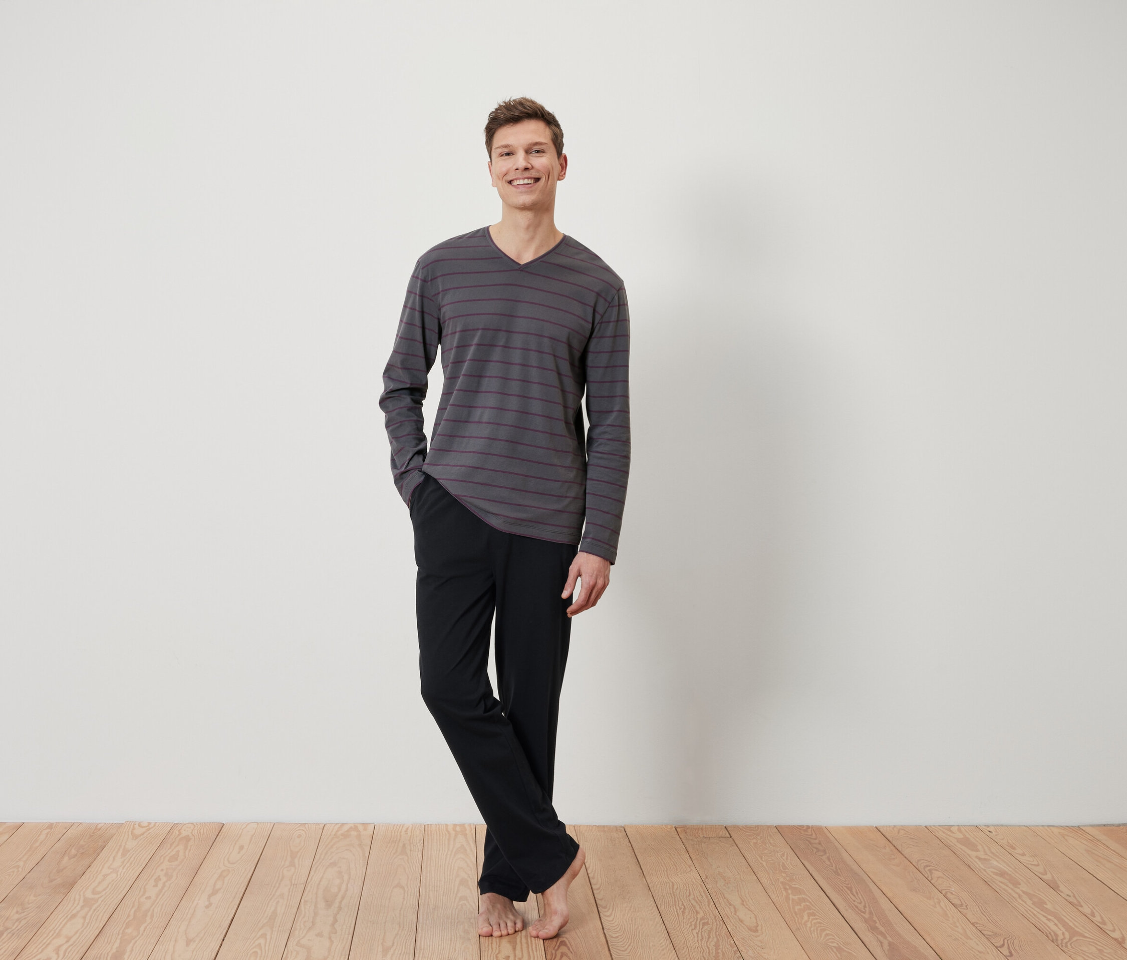 Un homme se tient pieds nus sur un plancher en bois, vêtu d'un t-shirt à manches longues rayé gris et d'un pantalon noir.