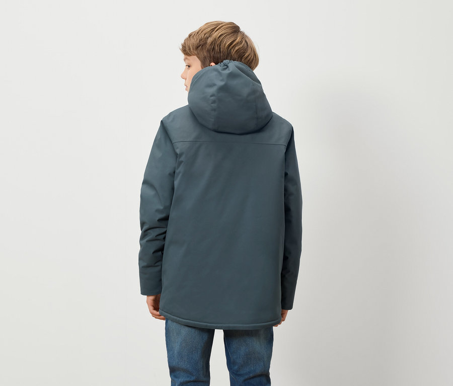 Un garçon vêtu d'une veste à capuche bleue et d'un jean bleu se tient dos à l'appareil photo devant un fond blanc.