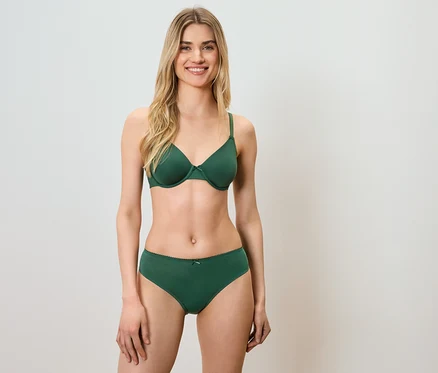 Une femme en soutien-gorge et culotte verts se tient devant un fond blanc et sourit.
