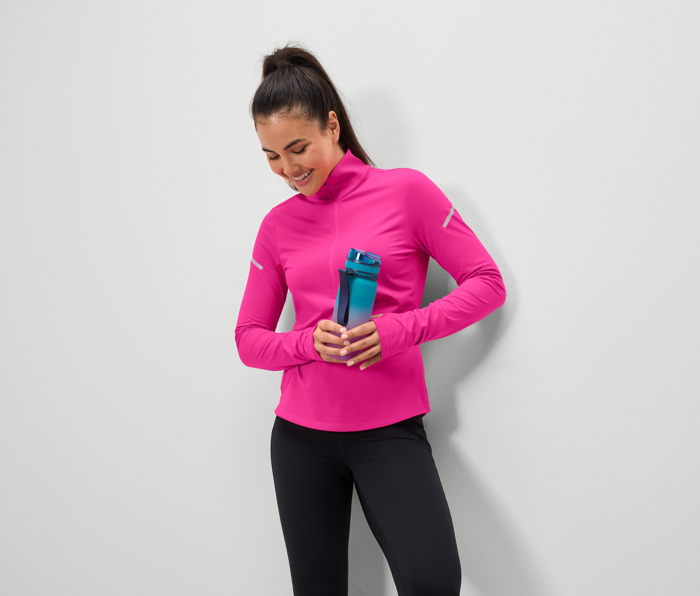 Une femme souriante tient une bouteille d'eau bleu-vert, porte un haut rose et un legging noir.