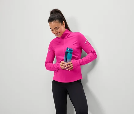 Une femme souriante tient une bouteille d'eau bleu-vert, porte un haut rose et un legging noir.
