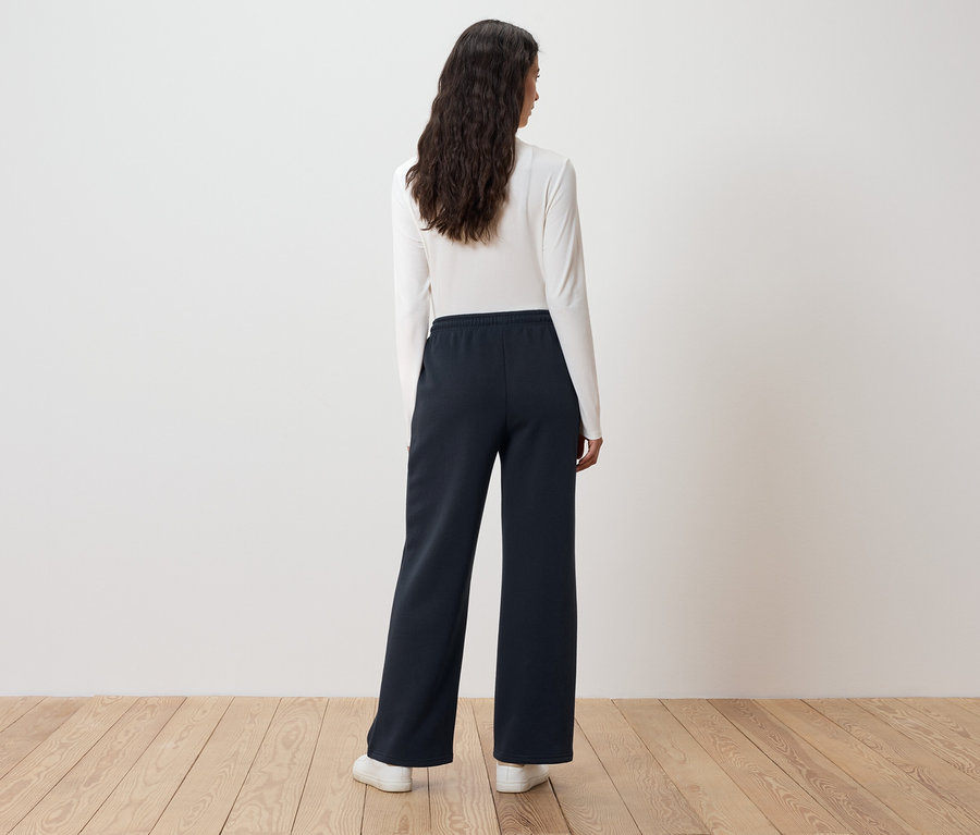 Une femme aux longs cheveux ondulés se tient dos à la caméra et porte un haut blanc à manches longues et un pantalon large bleu foncé.
