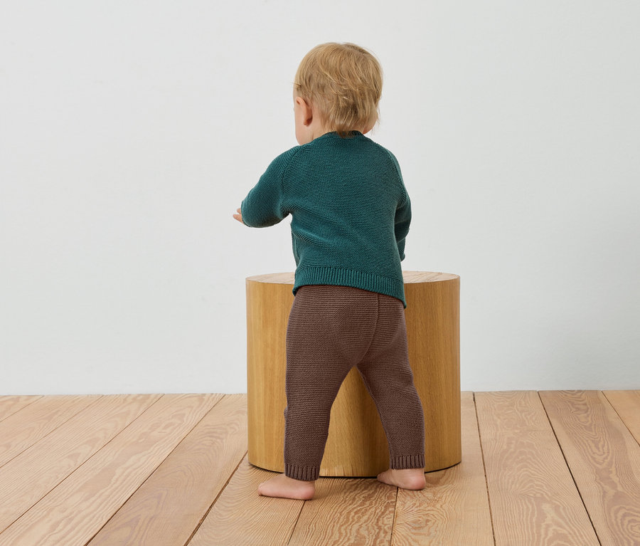 Un bébé portant un pull vert et un legging marron se tient près d'un tabouret en bois.