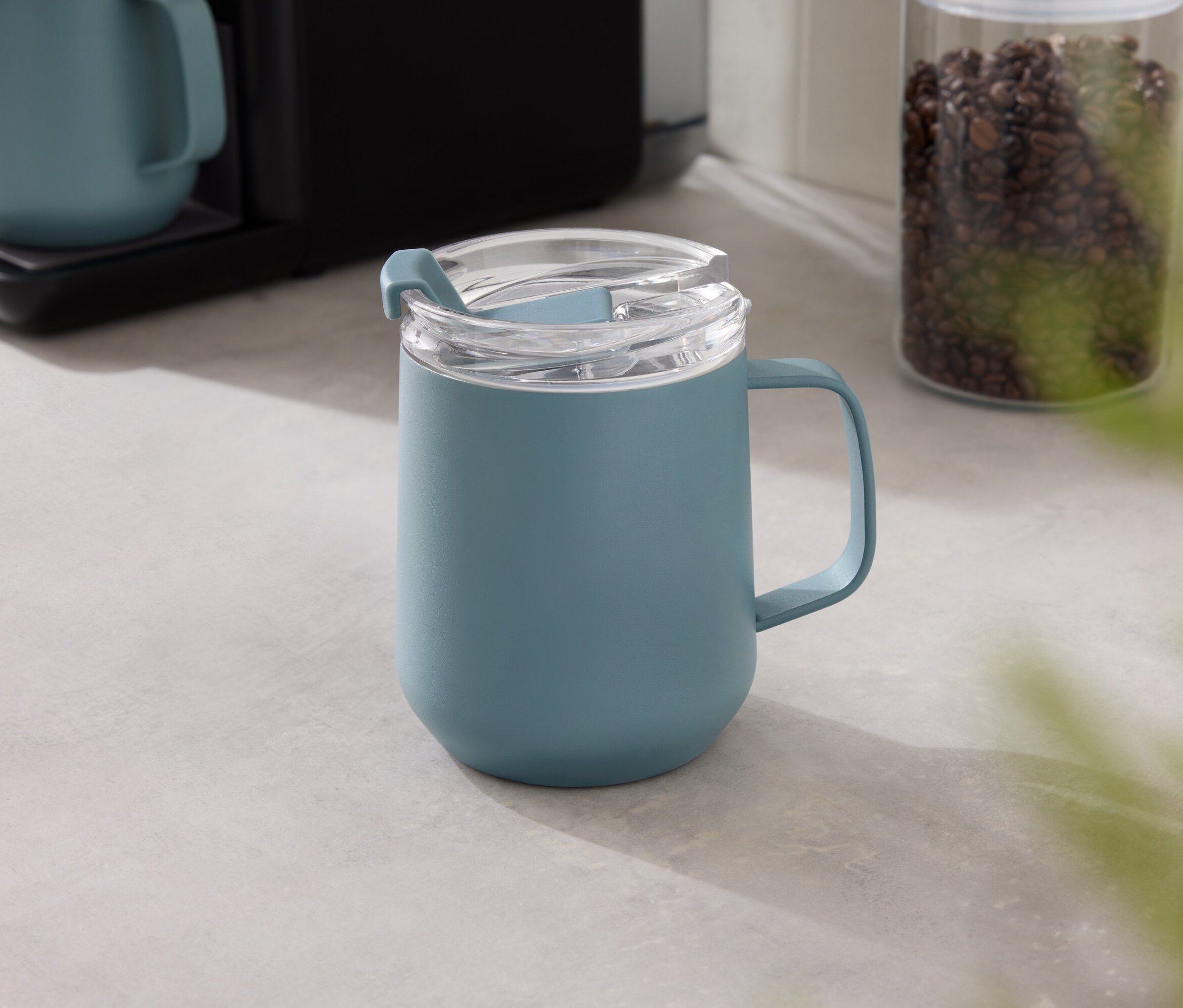 Une tasse bleue avec un couvercle transparent est posée sur une table grise, avec une machine à café et un récipient de grains de café en arrière-plan.