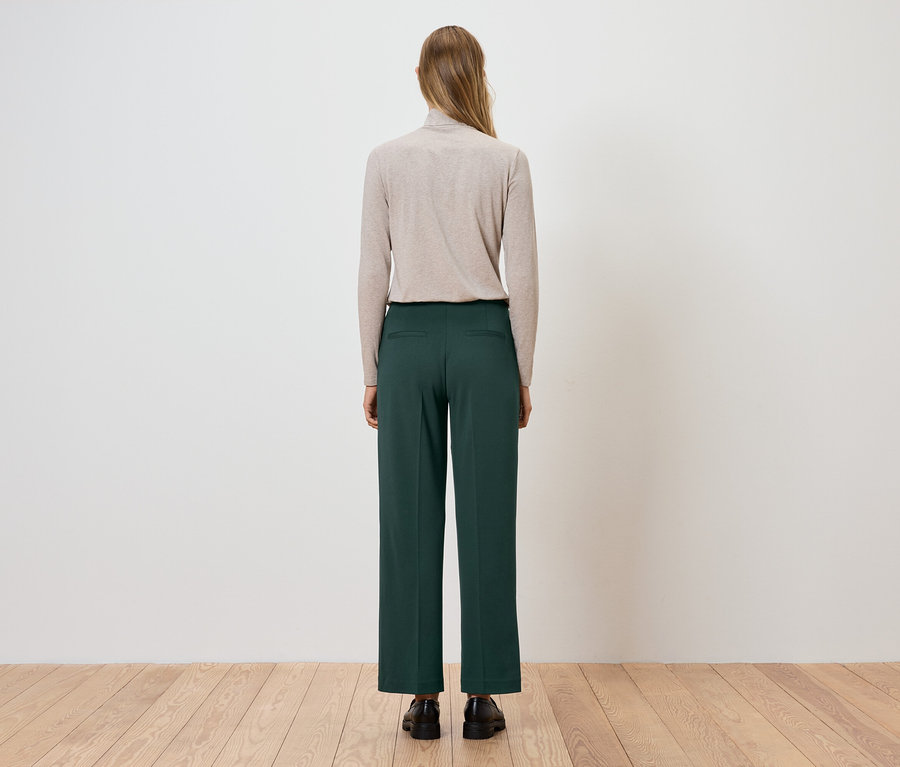 Une femme se tient dos à la caméra, portant un pull à col roulé beige et un pantalon vert.